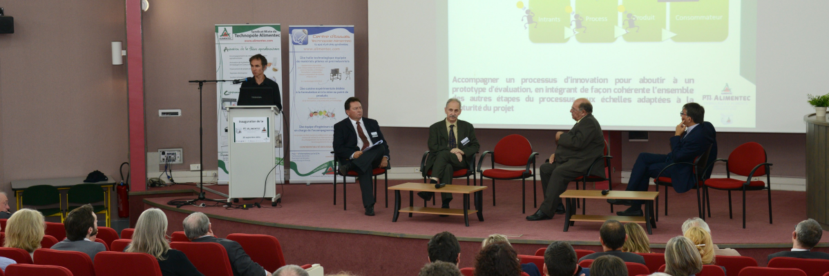 Salle de conférence avec scènes, podium, projecteur, écran, cinq personnes assises sur scène, public assis.