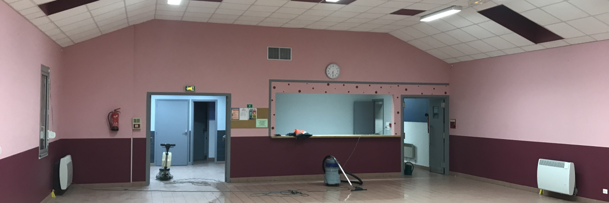 Salle avec sol carrelé, murs partagés rose et marron, plafond voûté, éclairage fluorescent, porte, fenêtre, comptoir, horloge, radiateurs et balai.