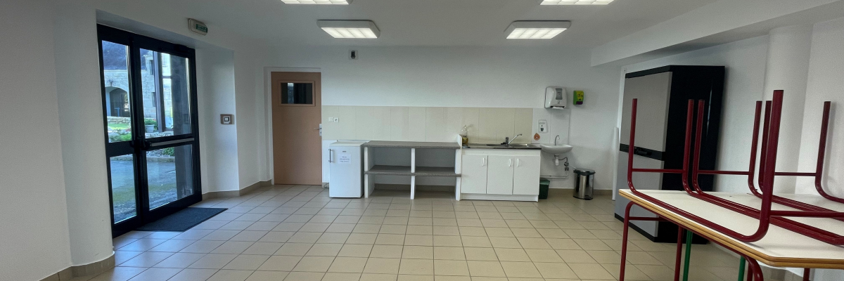 Salle avec tables, chaises, évier, réfrigérateur, et porte. Sol carrelé, lumières au plafond.