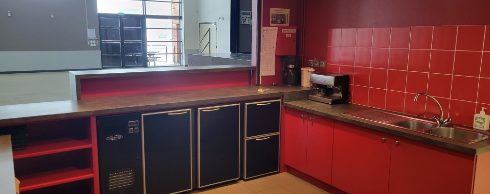 Cuisine avec plans de travail, armoires, évier, réfrigérateur, cafetière et étagères.