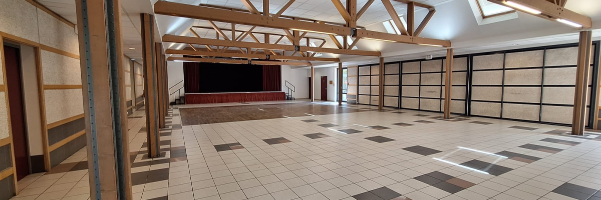 Salle spacieuse avec piliers en bois, sol carrelé, escaliers, portes vitrées et plafond haut.