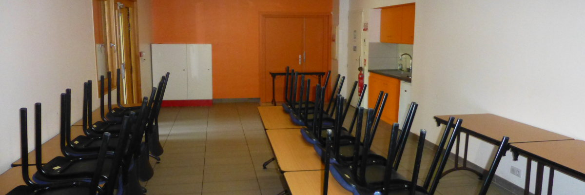 Salle avec tables, chaises, tableau et équipement électrique. Murs orange, sol gris et éclairage intégré au plafond.