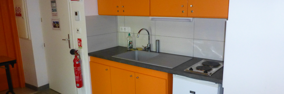 Cuisine avec plans de travail, évier, plaques de cuisson, armoires orange, frigo blanc, extincteur et signalétique de sécurité.