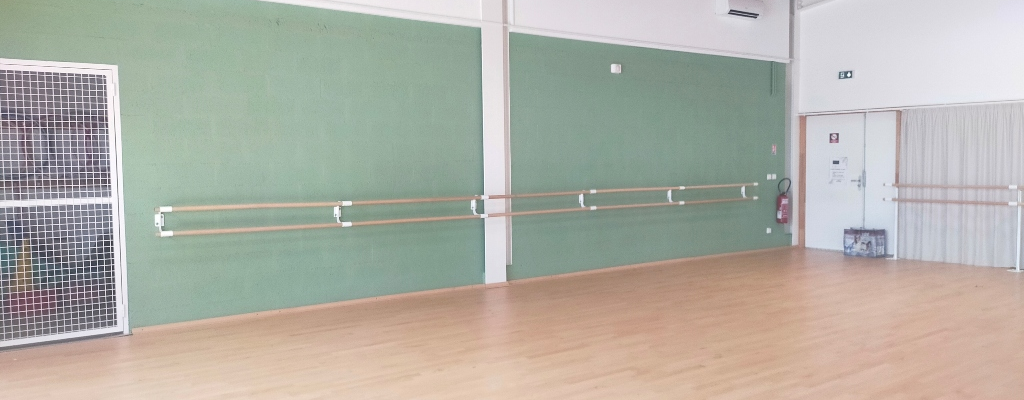 Salle avec barres de danse, mur vert, parquet, porte, extincteur et ventilateur.