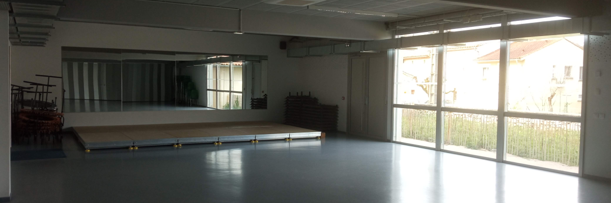 Salle vide avec miroir, plateforme, chaises empilées, grandes fenêtres et éclairage.