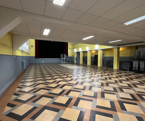 Grande salle avec motif de sol carrelé, éclairage au plafond, murs jaunes et porte au fond.