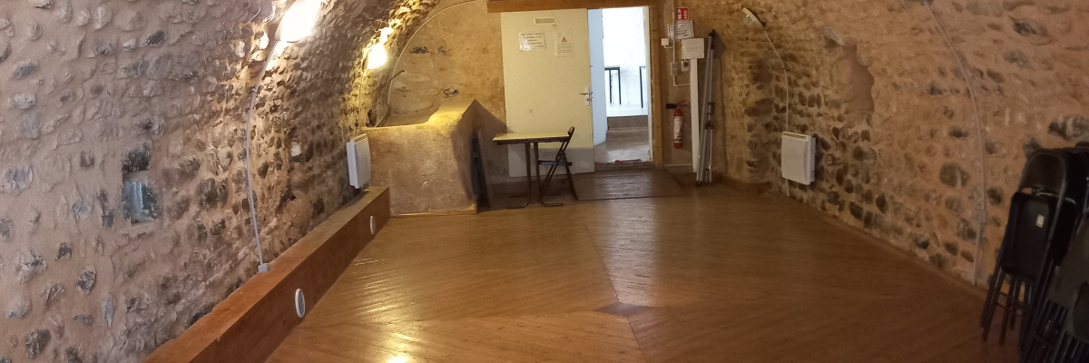 Pièce voûtée avec plancher en bois, fenêtres étroites, lustre, meubles et chaises.