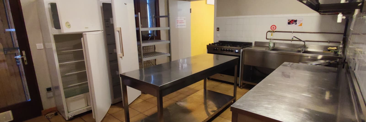 Cuisine avec réfrigérateur, étagères, tables en acier inoxydable, évier et porte jaune.