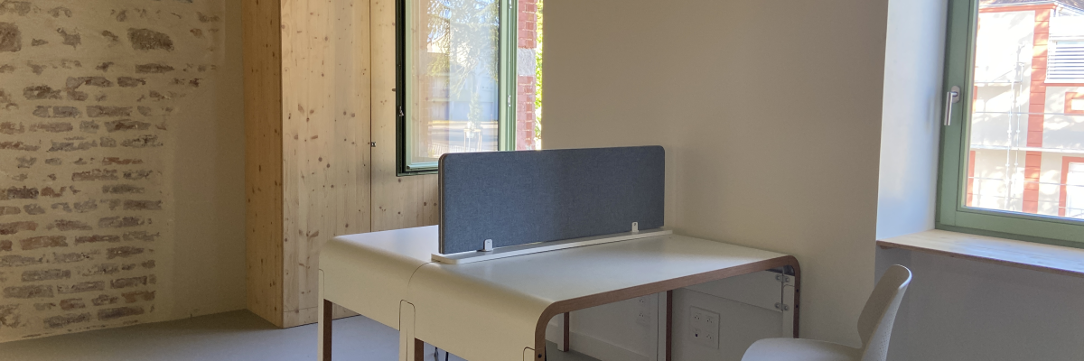Bureau avec séparateur, chaise et tabouret près de fenêtres. Mur en brique et armoire en bois.