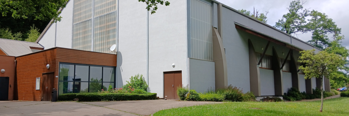 Bâtiment avec façade principale blanche, toiture grise, et extension en brique. Grandes fenêtres, porte centrale, pelouse verte et allée.