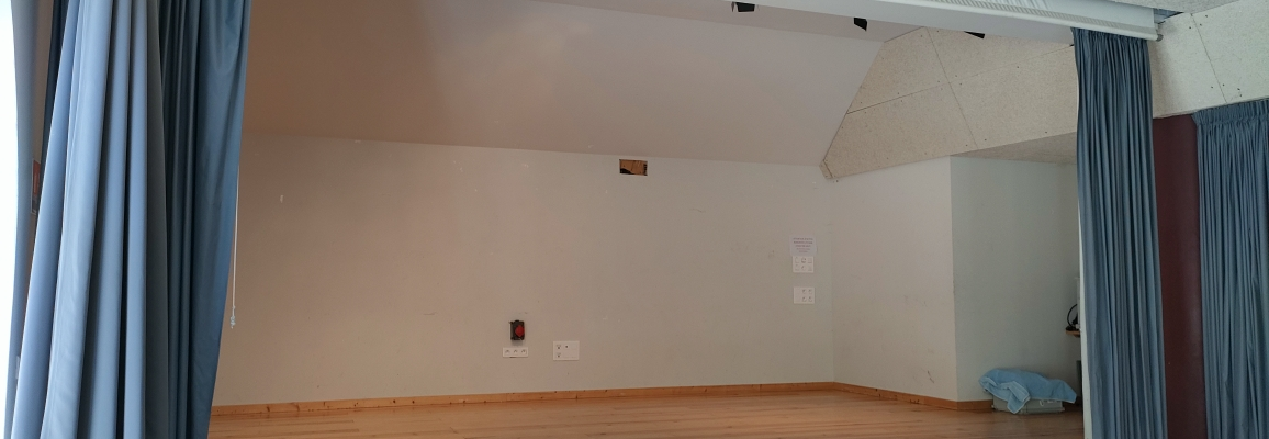 Salle avec scène en bois, rideaux, projecteur, prise électrique et escalier.