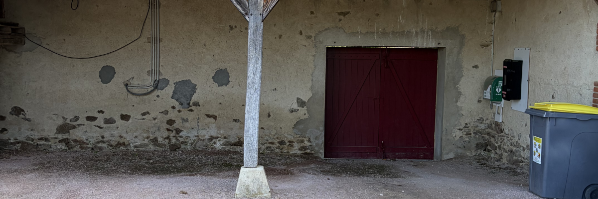 Bâtiment avec toit en tuiles, porte rouge, poubelle jaune et sol en gravier.