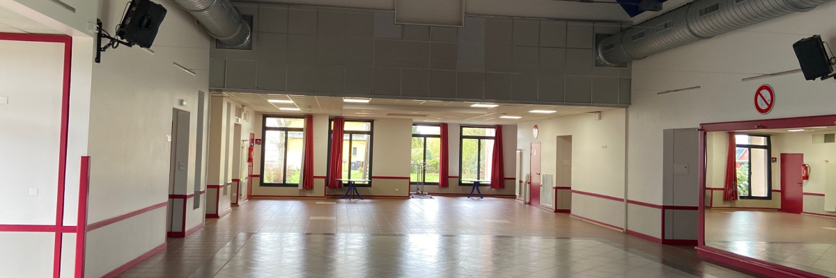 Grande salle vide avec sols carrelés, murs blancs, cadre de fenêtres et de portes rouges, plafond bleu avec conduits et spots.