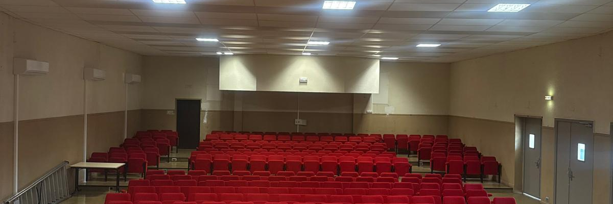 Salle avec rangées de sièges rouges, scène en bois, éclairage au plafond.
