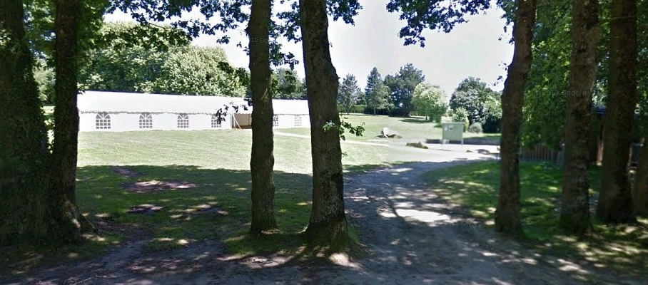 "Allée de gravier entre des arbres menant à un bâtiment blanc dans un parc avec pelouse et végétation."