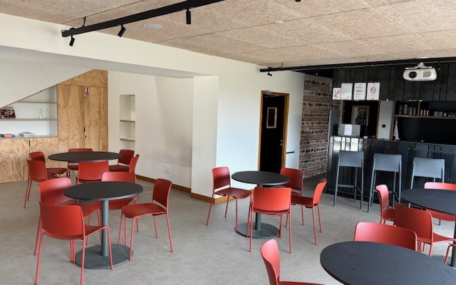 Salle avec tables rondes, chaises rouges, cuisine et projecteur.