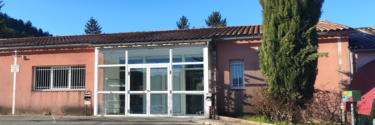 Bâtiment rose avec toiture en tuiles, portes vitrées et fenêtres grillagées. Arbre et panneau de stationnement visible.