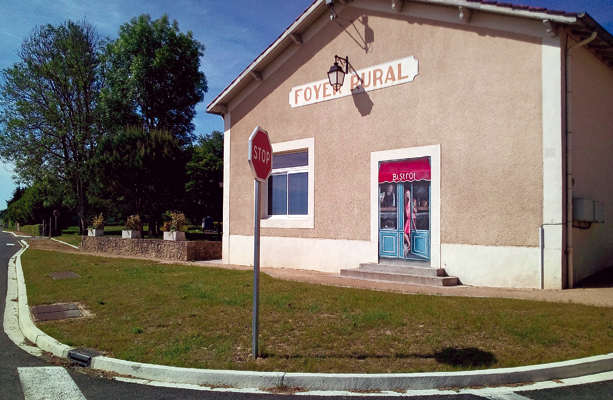 Bâtiment avec enseigne "Foyer Rural", porte avec enseigne, panneau stop, arbre, route et pelouse.