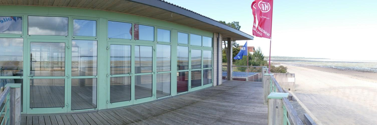 Bâtiment avec terrasse donnant sur une plage, porte vitrée, rampes, et drapeau.