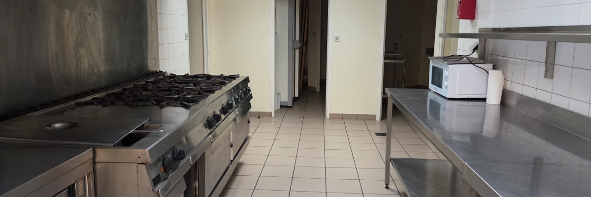 Cuisine avec plans de travail, fourneaux, hottes, frigo et micro-ondes. Sol carrelé, murs blancs. Porte et poubelle visibles.