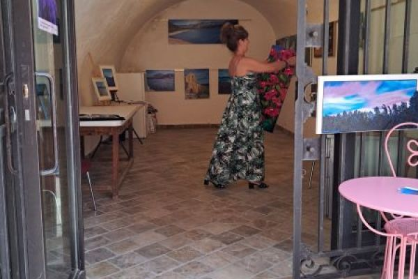 Galerie avec tableaux, écrans, tables, chaises. Personnes en arrière-plan.