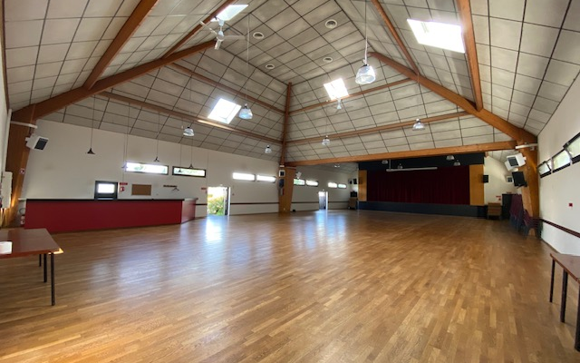 Salle avec parquet, plafond haut, tables, chaises, rideaux, scène et éclairage.