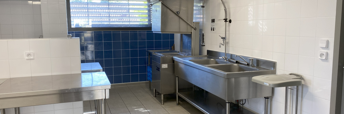 Cuisine professionnelle avec plans de travail, évier, rinçage, chariot de service, hottes et carrelage bleu et blanc.