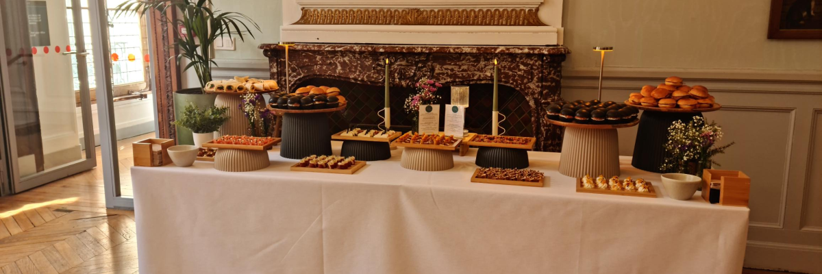 Buffet avec divers plateaux de nourriture, nappage blanc, miroir au-dessus, plantes, lustres, porte vitrée.