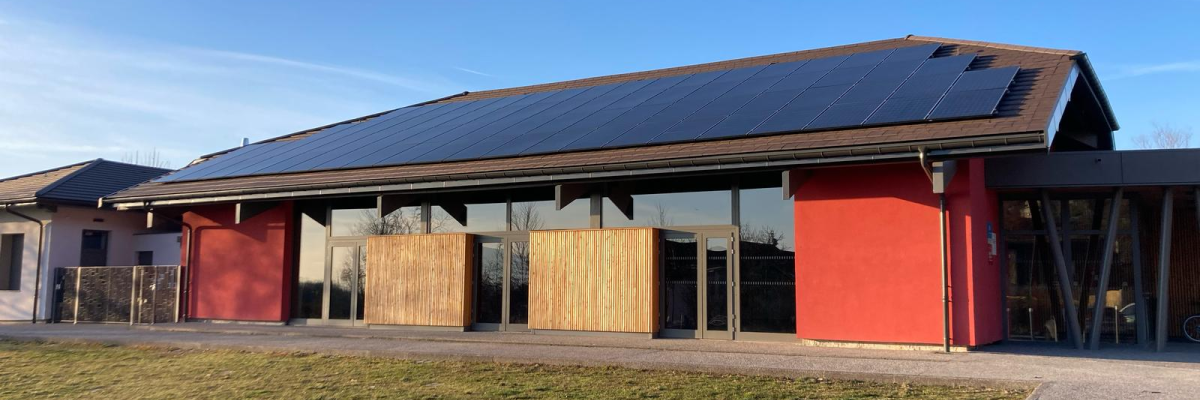Bâtiment avec toiture en panneaux solaires, murs rouges et ouvertures en bois. Pelouse et allée en gravier.
