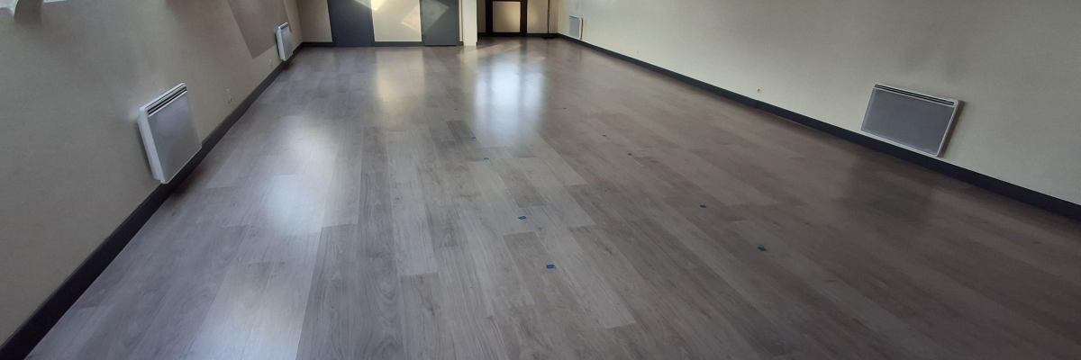 Salle vide avec parquet, fenêtres, radiateurs et éclairages encastrés.