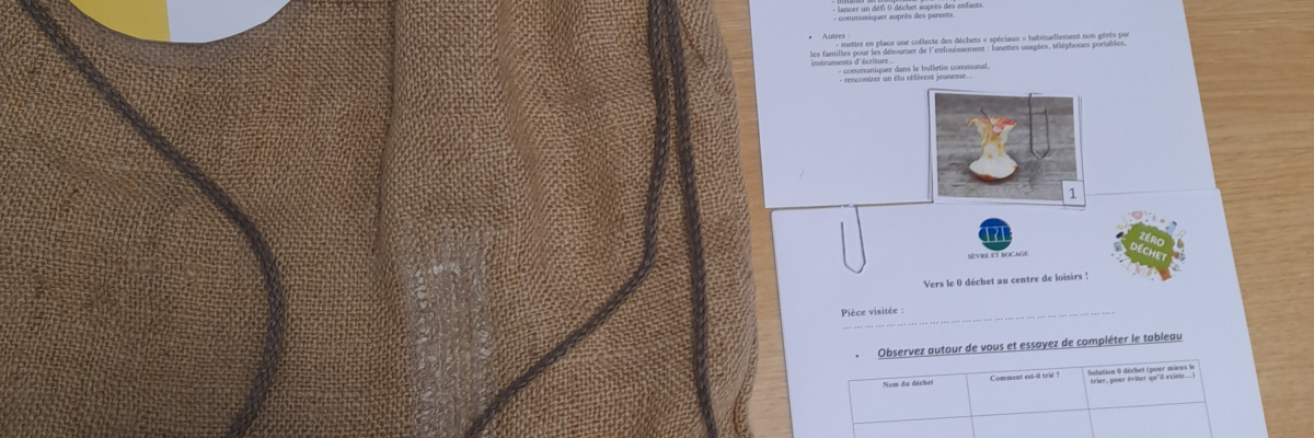 Sur une table, un sac en toile avec une étiquette, une feuille de papier avec du texte et une image, et un petit objet gris.