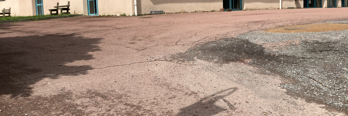 Bâtiment avec portes, fenêtres, tables et bancs. Ciel couvert. Sol en gravier et terre.