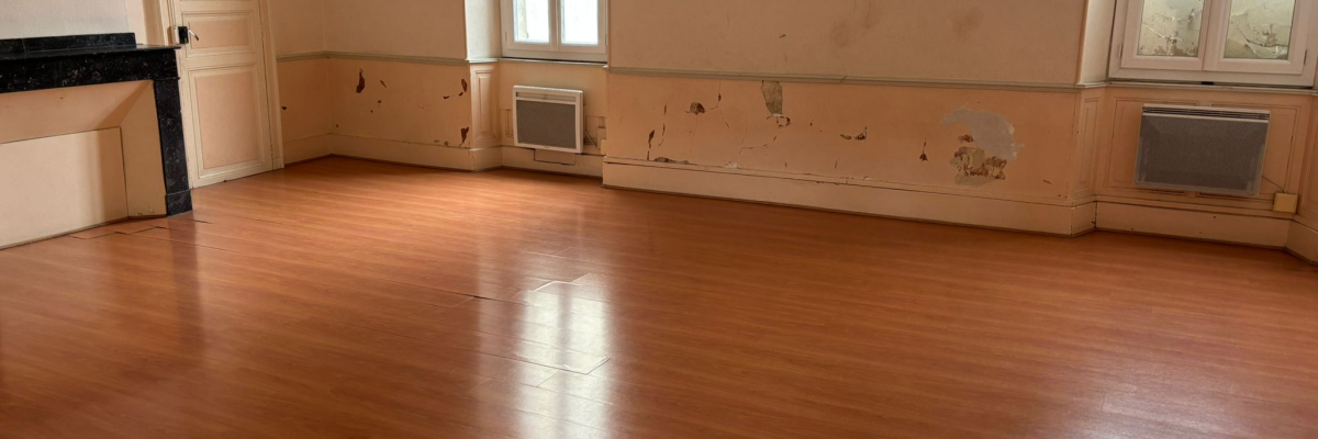 Pièce vide avec parquet, murs endommagés, radiateurs et fenêtres.