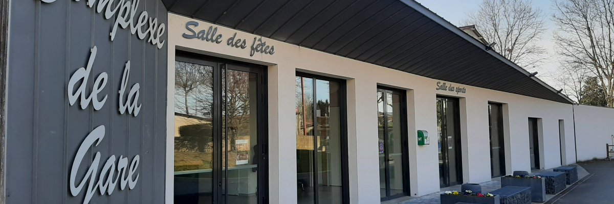 Bâtiment moderne avec des portes vitrées, des murs gris et blancs, une enseigne "Complexe de la Gare" et des jardinières.