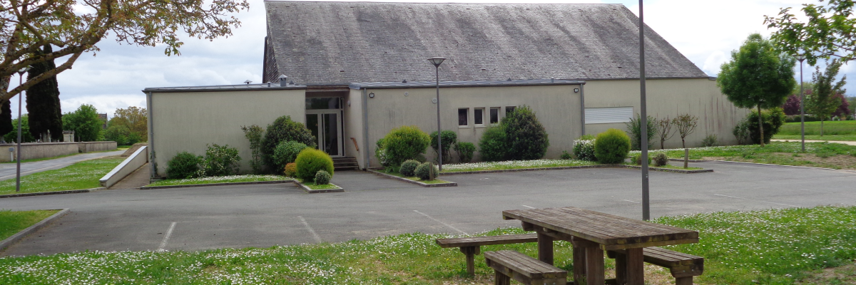 Bâtiment avec toiture grise, entrée, jardins, parking, lampadaires et banc en bois.