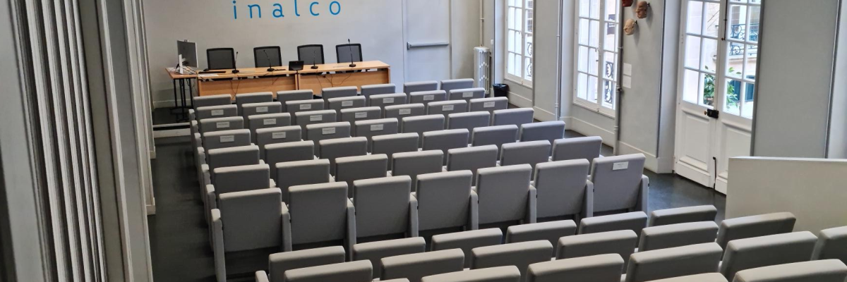 Salle de conférence avec rangées de chaises, table, projecteur, écran et horloge.