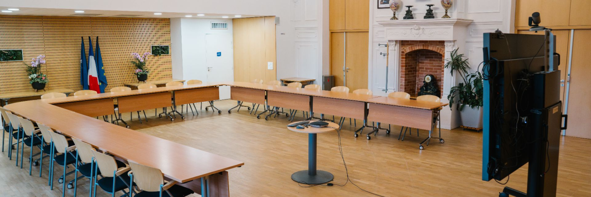 Salle avec tables, chaises, écran, pied de micro, cheminée, statue, porte, plantes.