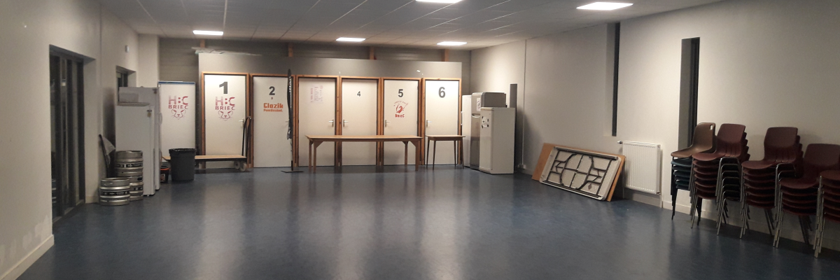 Salle vide avec tables, chaises, portes numérotées, radiateurs et frigo.