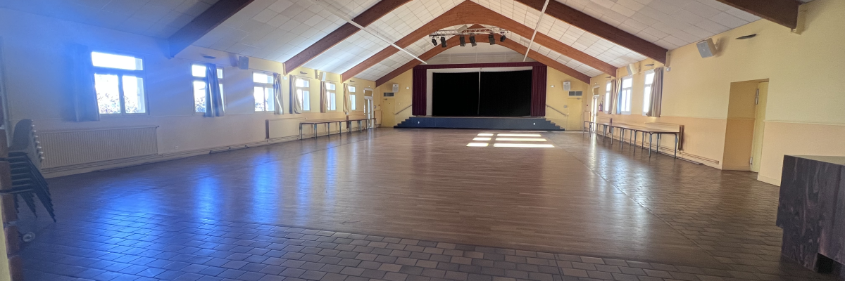 Grande salle avec sol en bois, murs blancs, plafond voûté, fenêtres, rideaux, radiateurs et scène.
