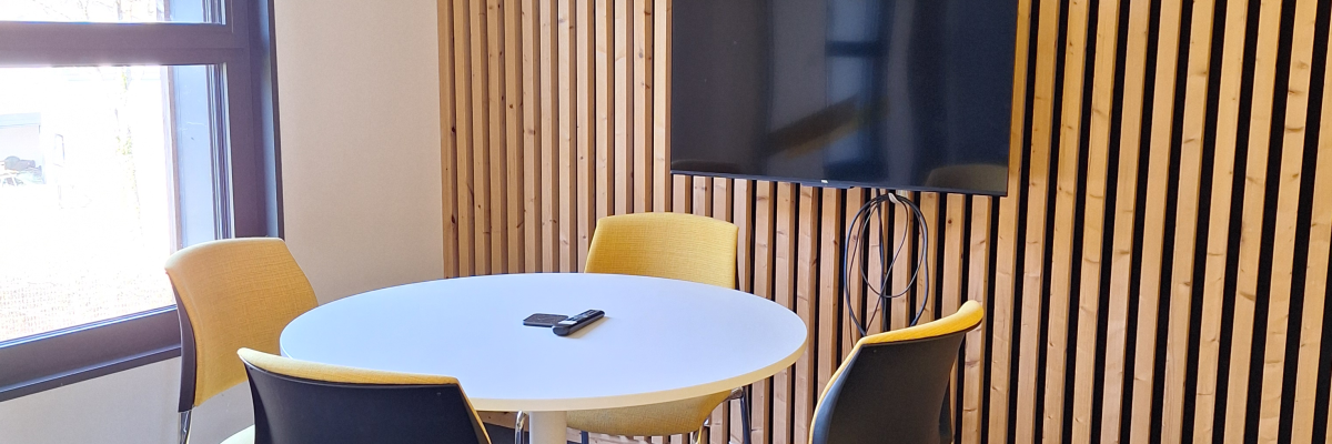 Table ronde avec chaises, téléviseur sur support, mur à rayures verticales.
