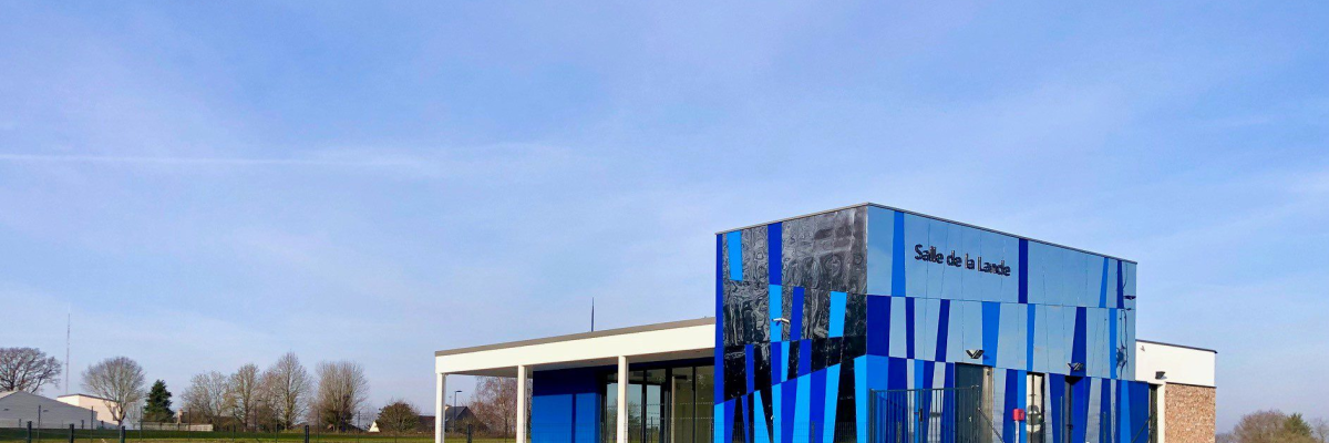 Bâtiment moderne avec façade bleue et blanche, entouré de terrain herbeux et clôturé.