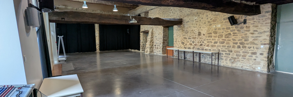 Grande salle avec sol en bois, murs en pierre, plafond à poutres apparentes, comptoir, tables, projecteurs et éclairages suspendus.