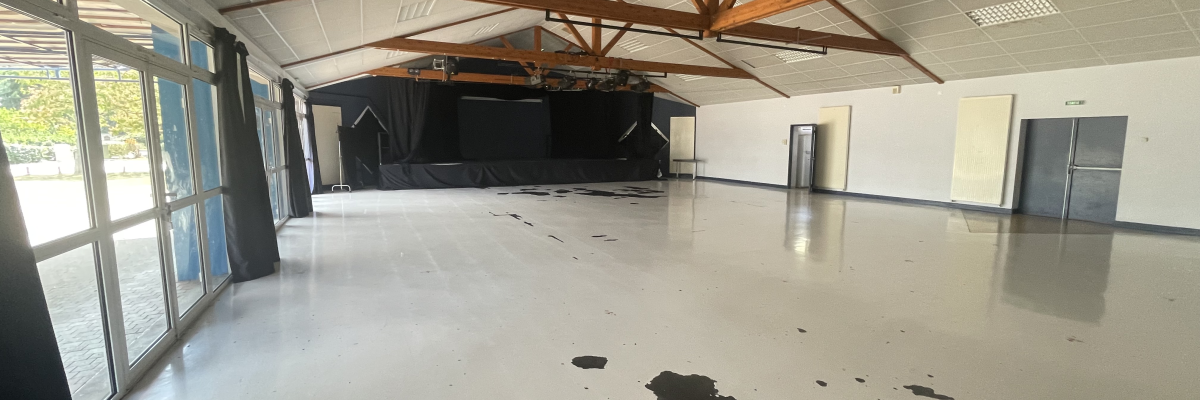 Salle vide avec sol blanc, poutres apparentes, portes et taches noires au sol.