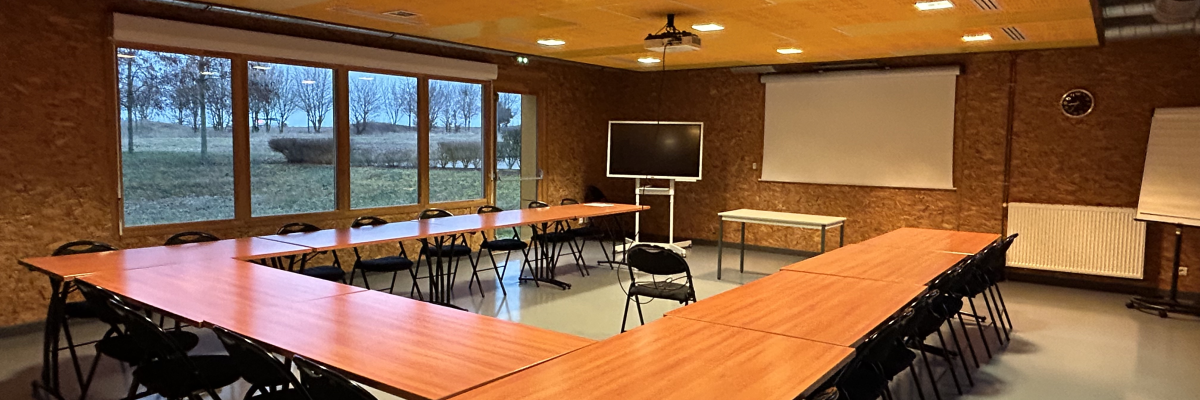 Salle avec longues tables, chaises, écran, tableau blanc et radiateur. Grandes fenêtres.