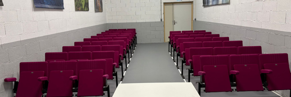 Salle avec rangées de chaises, table au centre, tableaux aux murs, porte.