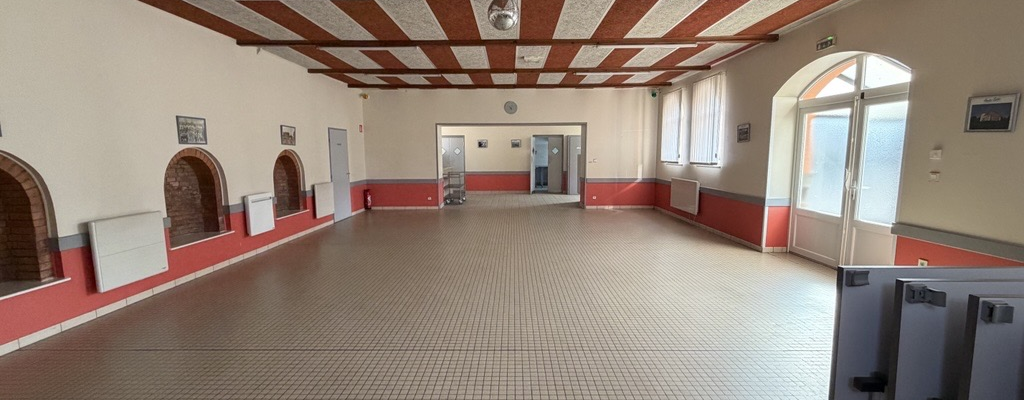 Salle vide avec sol carrelé, plafond à poutres apparentes, portes et fenêtres.
