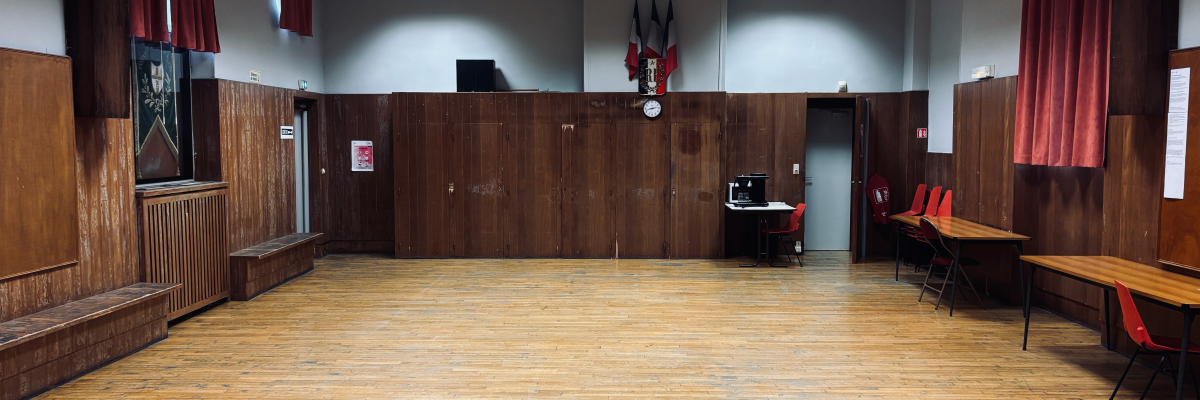 Salle avec parquet, bancs, tables, chaises, rideaux, porte, horloge et tableau.