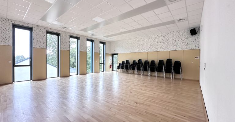 Salle spacieuse avec parquet, chaises alignées, grandes fenêtres et murs en bois clair.