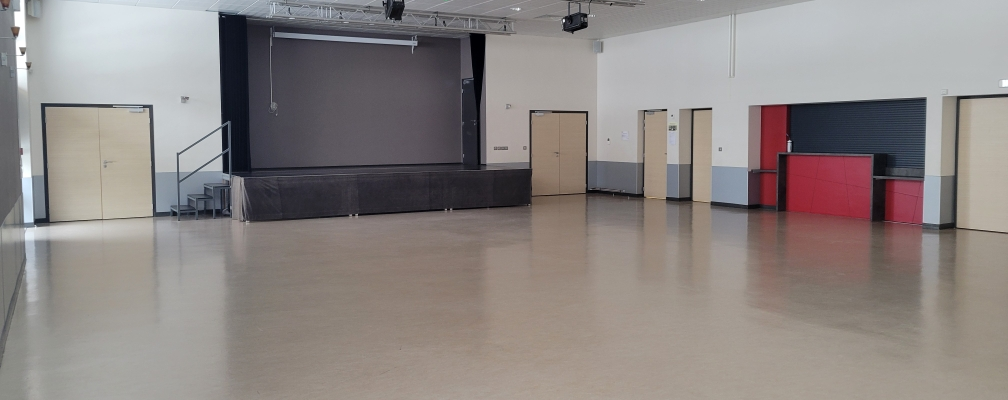 Salle spacieuse avec scène, écran, projecteurs, portes et comptoir rouge.