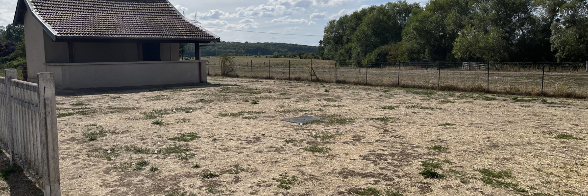 Bâtiment avec toit en tuiles, terrain sec, clôture en bois, arbres en arrière-plan.